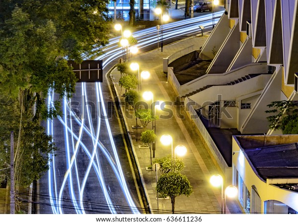 Light trail of cars making the curve of a\
street of a city. View from above, sidewalk, post lights, some\
trees around and\
buildings.