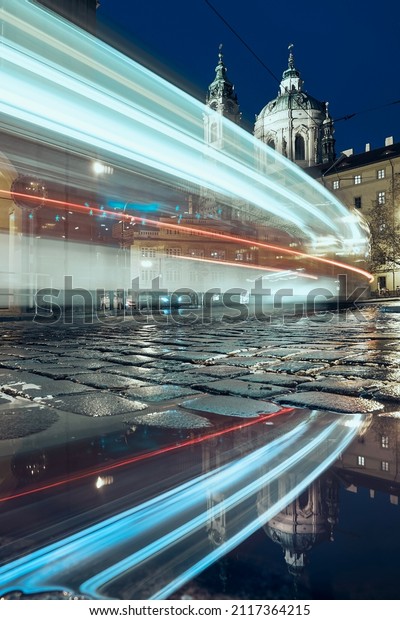 Light trail of tram passing between historical\
buildings. Reflection in puddle on wet road. Lesser Town in Prague,\
Czech Republic\
