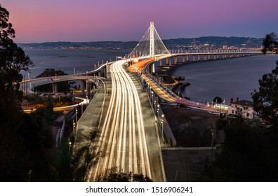 The light trails of rush hour traffic on the Bay Bridge facing towards Berkeley and Oakland after the sun has set and the sky is a deep pink, from Treasure Island, San Francisco, USA
