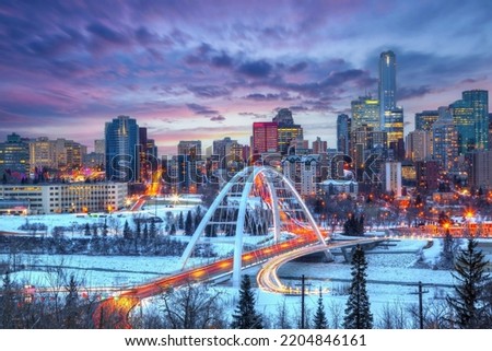 Light trails from rush hour traffic lit up Edmonton downtown Winter sunset skyline showing Walterdale Bridge across the frozen, snow-covered Saskatchewan River and surrounding skyscrapers.