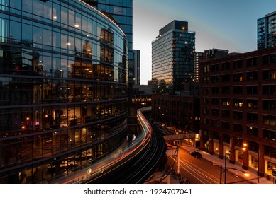 Light Trails of a train passing through Chicago City with Buildings standing all around it and skyline reflecting off building big glass windows at night 