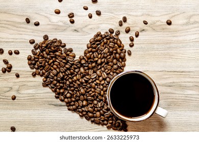 A light wooden table with roasted coffee beans arranged in a heart shape in the center, surrounded by scattered beans, and a white cup of black coffee placed near the heart. 