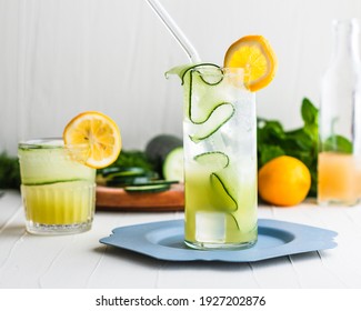 Light Yellow Green Gin Cocktails In A Highball Glass And Rocks Glass Surrounded And Garnished With Cucumbers And Lemon.