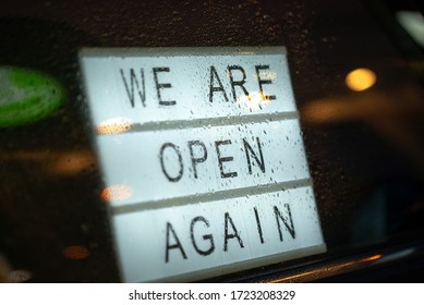 Lightbox with a sign we are open again behind a glass door of the cafe during the rain in the evening. We're open again after quarantine, photo of small business owner. Please wear a face mask and