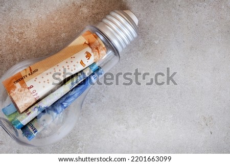 Lightbulb filled with money, South African Rand on mottled grey and selective focus