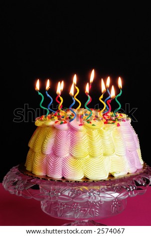Lighted Birthday Cake - Lighted candles on a pink and yellow iced birthday cake on a decorative cake plate in front of a black background.