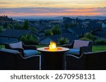 A lighted fire pit and patio set overlooking a suburb of modern homes with the city skyline behind at early evening.