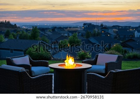 A lighted fire pit and patio set overlooking a suburb of modern homes with the city skyline behind at early evening.