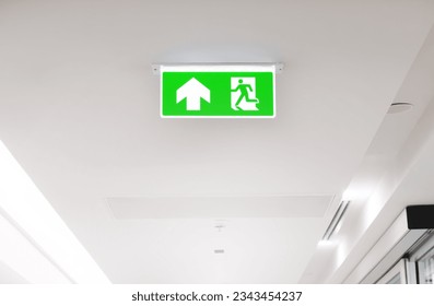 Lighted glowing green emergency exit signs in an office hallway with arrows pointing the way out of the building. - Powered by Shutterstock