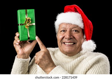 Light-hearted Male Senior With A Red Father Christmas Cap Cracking A Smile. He Is Pointing At A Long, Green, Wrapped Gift Raised Above Eye-level In His Right Hand. Golden Bowknot. Isolated On Black.