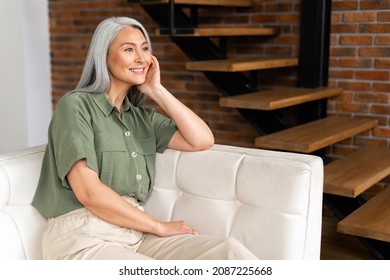 Light-hearted Positive Middle-aged Woman With Long Gray Hair Sitting On Sofa In Modern Apartment, 50s Multiracial Asian Female Resting At Home, Looks Away Lost In Dreams, Wellbeing Concept