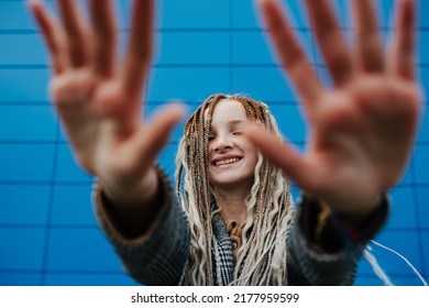 Lighthearted teenage girl blocking camera with hands in front of a blue panel wall covering. She is wearing a grey checkered jacket. - Shutterstock ID 2177959599
