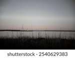 Lighthouse across calm water with tall marsh grass in the foreground, under a soft sky at dusk.