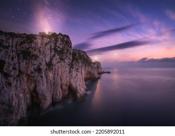 Lighthouse on the mountain peak at starry night in summer. Beautiful cliffs, rocky sea coast, bright stars, milky way and violet sky with clouds at dusk. Lighthouse of Cape Lefkada, Greece. Landscape - Shutterstock ID 2205892011