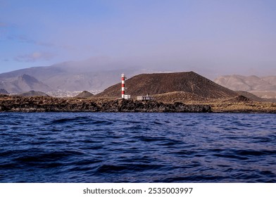 A lighthouse is on a rocky island in the ocean. The water is calm and the sky is clear - Powered by Shutterstock