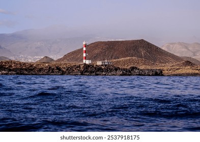 A lighthouse is on a rocky island in the ocean. The water is calm and the sky is clear - Powered by Shutterstock