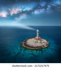 Lighthouse on smal island in the sea and Milky Way at night in summer. Aerial view of beautiful lighthouse with light beam on the rock, blue sky with stars. Landscape. Adriatic sea, Croatia. Space