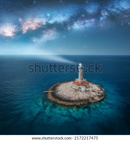 Lighthouse on smal island in the sea and Milky Way at night in summer. Aerial view of beautiful lighthouse with light beam on the rock, blue sky with stars. Landscape. Adriatic sea, Croatia. Space