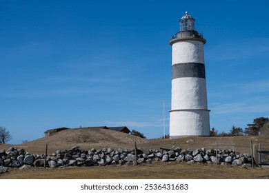 The lighthouse, painted in classic black and white stripes, overlooks a peaceful landscape. The sunlit day enhances the natural beauty of the rocky surroundings and grassy hill - Powered by Shutterstock