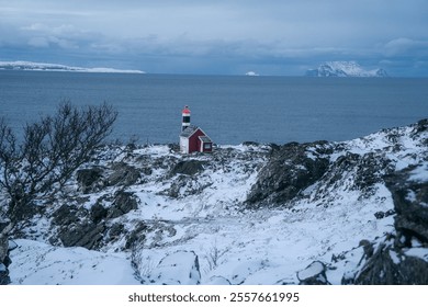 Lighthouse and a red cottage stand on a snowy, rocky coastline under a serene blue twilight. A distant boat sails on calm waters with snow-capped mountains in the background, creating a tranquil  - Powered by Shutterstock