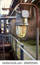 Lighting mast and selective focus explosion-proof and fireproof design rusty retro style lantern closeup over background of pipelines buildings and equipment of chemical plant with copy space.