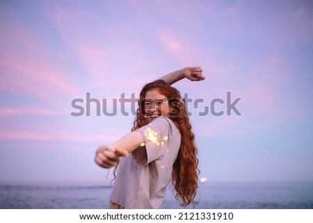 Lighting things up at the beach. Happy young woman dancing and laughing while holding a sparkling light in her hand. Cheerful young woman celebrating with bengal lights at the beach.