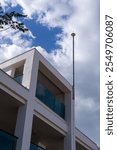 A lightning rod. A contemporary building features sleek white design and glass balconies. A tall antenna rises above the building, set against a backdrop of dynamic clouds on a clear day.