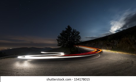 Lights On The Asphalt, At Night On A Mountain Road, Illuminated By The Moon Begins To Rise Over The Horizon, A Car Making A Turn, Leaving Behind A Trail Of Light.