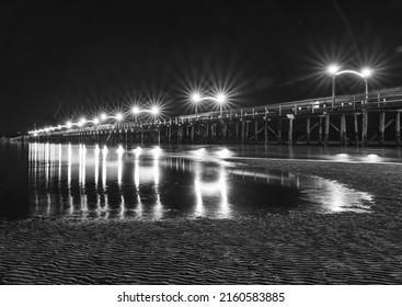 The lights of Pier at night reflected in the wet sand on the beach. Wooden pier at White Rock, BC extends diagonally into image. Black and white photo. Nobody, selective focus
