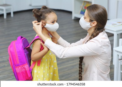 A Light-skinned Teacher In A Medical Mask For Respiratory Protection Puts A Mask On The Face Of A Dark-skinned Student With A Backpack On Her Shoulders.