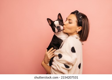 Light-skinned Young Brunette Woman Kisses Her Pet Boston Terrier Hard On Pink Background. Girl In White Sweater Loves Dogs Very Much. True Friendship, Wonderful Happy Moments