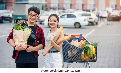 Likable Asian Couple Hold Paper Eco Bags With Organic Healthy Food In Hands While Standing Near Store Mall. Happy Family Shopping On Weekend.