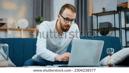 Likable concentrated skilled 30-aged bearded man sitting on the couch at home and typing on computer comparing datas with financial reports