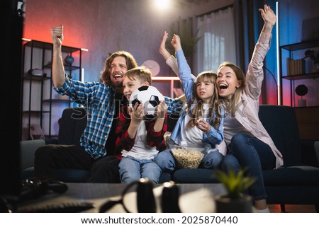 Likable excited family of four, football fans sitting on sofa in front of TV at home in the evening and celebrating winning and scored goal with raised hands and shouts