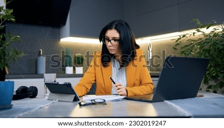 Likable high-skilled modern 35-aged dark-haired businesswoman in formal clothes sitting at home on the kitchen and revisioning documents and comparing with datas on tablet pc,front view