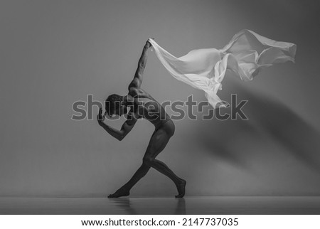 Like ancient sculpture. Black and white portrait of graceful muscled male ballet dancer dancing with fabric, cloth isolated on grey studio background. Grace, art, beauty, contemp dance concept.