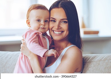 Like Mother Like Daughter. Cheerful Beautiful Young Woman Holding Baby Girl In Her Hands And Looking At Camera With Smile While Sitting On The Couch At Home