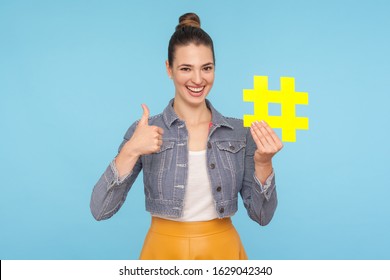 Like to popular blog and trends. Joyful attractive woman with hair bun in stylish outfit holding big yellow hashtag symbol and showing thumbs up gesture. indoor studio shot isolated on blue background