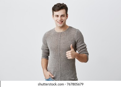 I Like That. Good Job. Happy Caucasian Male With Dark Hair Wearing Casual Long Sleeved Sweater Making Thumbs Up Sign And Smiling Cheerfully, Showing Support And Respect To Someone. Body Language