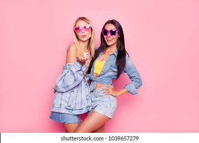 I like you! Beautiful playful cheerful women dressed in fashionable stylish shorts, shirt, jacket, top, funny star and heart glasses are embracing, sending air-kiss, isolated on bright pink background