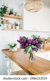 Lilac flowers in vase standing on wooden countertop in the kitchen. Modern white u-shaped kitchen in scandinavian style. Open shelves in the kitchen with plants and jars.