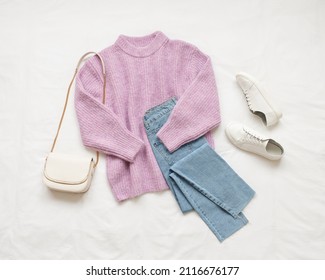 Lilac Knitted Sweater, Blue Jeans, White Sneakers And Bag Lie On White Background. Overhead View Of Woman's Casual Outfit. Trendy Stylish Women Clothes. Flat Lay, Top View.