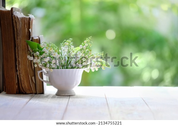 Lily of the valley flowers\
in cup and books on white table, green natural background. symbol\
of spring season. beautiful romantic floral composition. copy\
space