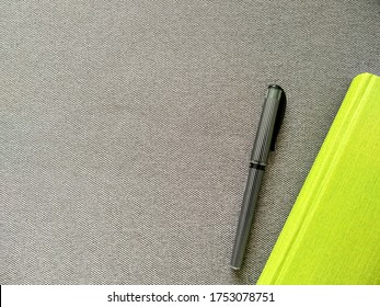 Lime Green Notebook With A Black Striped Pen On A Gray Textile Background With Space For Text. Diary For Planning Affairs On The Couch For Work. Bright Green On A Beautiful Gray.