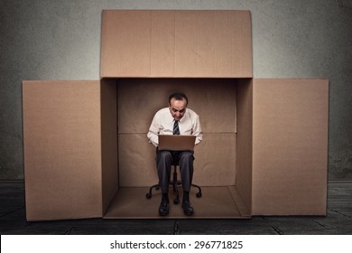 Limitations poor communication of corporate life. Portrait corporate middle aged man working on laptop sitting on chair inside carton box in empty office room