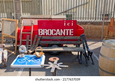 Limoux Aude France. 07.12.22 Bright Flea Market Scene, A Wooden Bench,  Red Brocante Sign With White Lettering. Cycle Path Icon Table. A Wine Barrel. Sach Window. Wall Lamp. Trailer And Iron Railings