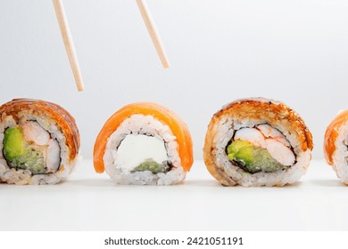 A line of assorted sushi rolls including Philadelphia and eel avocado rolls with a pair of chopsticks hover above a selection of sushi rolls, poised to pick up a delicious Philadelphia or eel avocado - Powered by Shutterstock