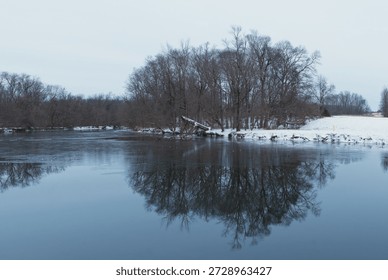 A line of bare leafless trees reflect off the surface of still water of a creek with snow covered shoreline on a overcast gloomy winter day after a snowstorm
