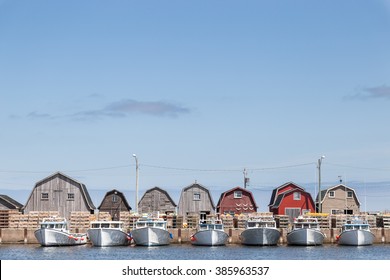 A line of fishing sheds and tied up fishing boats at Malpeque Harbour in Prince Edward Island (PEI) on the east coast of Canada. They sky is light blue and there are a few scattered clouds.