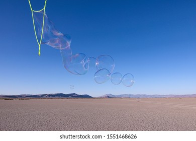 A line of a large soap bubbles fly away from long bubble wands, Alvord desert with Steens mountains in the background